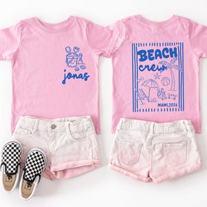 Op de afbeelding: Twee roze T-shirts met blauwe afbeeldingen. E&eacute;n heeft de naam "Jonas", de andere zegt "Beach Crew Miami, 2026" met strandillustraties. Ze worden getoond met witte shorts en zwart-wit geblokte instappers.
