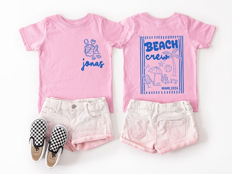 Op de afbeelding: Twee roze T-shirts met blauwe afbeeldingen. E&eacute;n heeft de naam "Jonas", de andere zegt "Beach Crew Miami, 2026" met strandillustraties. Ze worden getoond met witte shorts en zwart-wit geblokte instappers.