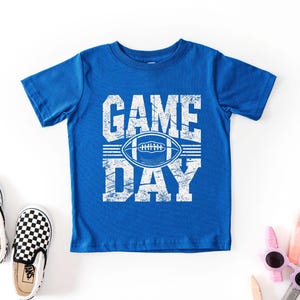 K&ouml;nnte beinhalten: Ein k&ouml;nigsblaues T-Shirt mit den Worten "GAME DAY" in wei&szlig;en Blockbuchstaben und einer wei&szlig;en Football-Grafik. Ein Paar schwarz-wei&szlig; karierte Slip-on-Sneaker und eine Sonnenbrille sind ebenfalls im Bild.