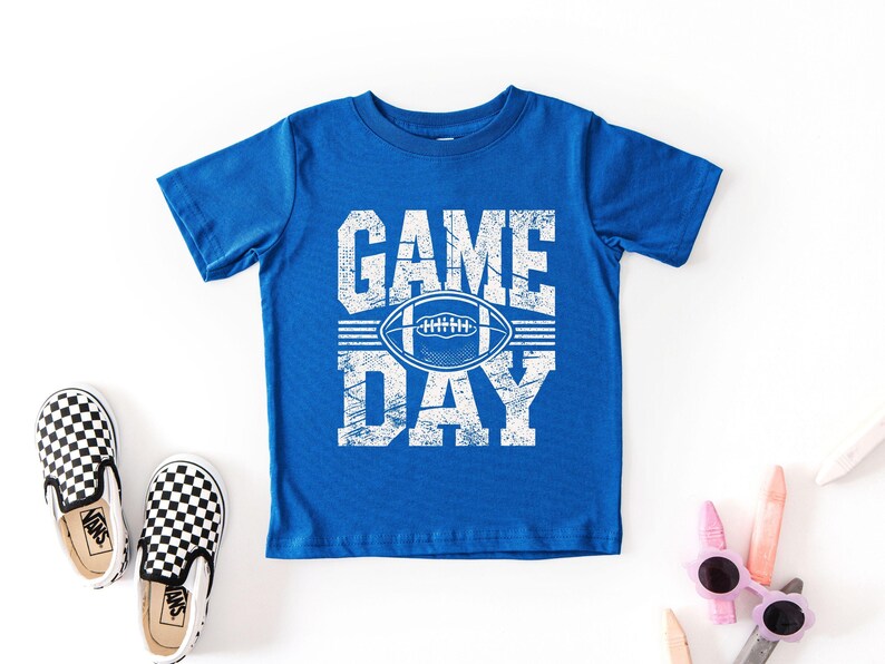 K&ouml;nnte beinhalten: Ein k&ouml;nigsblaues T-Shirt mit den Worten "GAME DAY" in wei&szlig;en Blockbuchstaben und einer wei&szlig;en Football-Grafik. Ein Paar schwarz-wei&szlig; karierte Slip-on-Sneaker und eine Sonnenbrille sind ebenfalls im Bild.