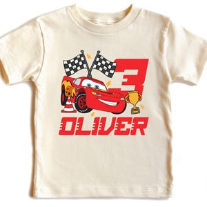 May include: Cream-colored t-shirt featuring a red cartoon race car, the number 3, and a gold trophy. The design includes checkered flags and the name "OLIVER" in red block letters. A fun, themed shirt.