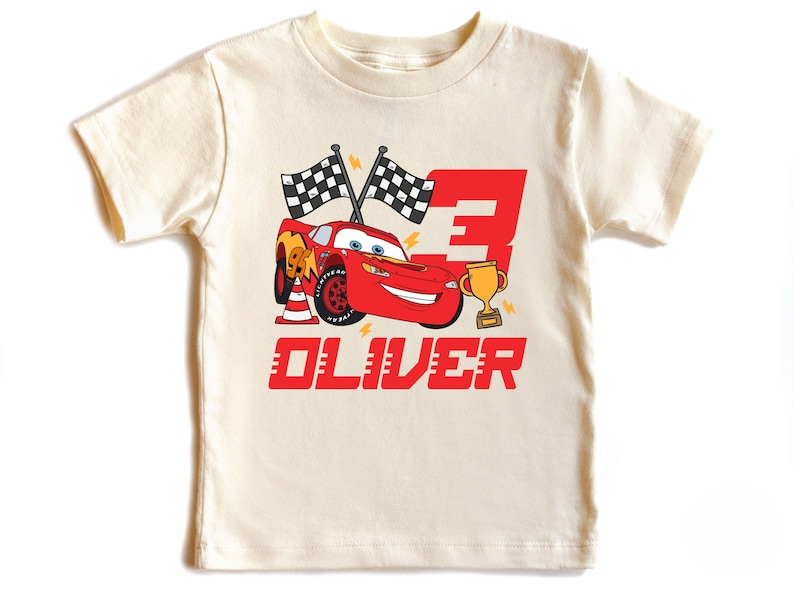 May include: Cream-colored t-shirt featuring a red cartoon race car, the number 3, and a gold trophy. The design includes checkered flags and the name "OLIVER" in red block letters. A fun, themed shirt.
