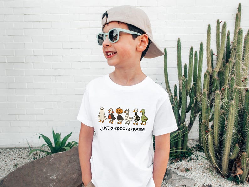 May include: White t-shirt with a Halloween-themed graphic of cartoon characters, including a ghost, vampire, pumpkin, mummy, skeleton, and goose. The text "just a spooky goose" is below the characters.