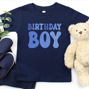 May include: A navy blue t-shirt with the text "BIRTHDAY BOY" in blue. The image includes navy blue slip-on shoes and trousers. A beige teddy bear is also present.