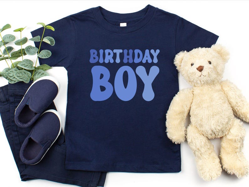 May include: A navy blue t-shirt with the text "BIRTHDAY BOY" in blue. The image includes navy blue slip-on shoes and trousers. A beige teddy bear is also present.
