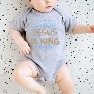 May include: A gray baby bodysuit with the text "JESUS IS KING" in gold and a light blue lion design. The baby is lying on a white surface with black speckles. The bodysuit is short-sleeved.