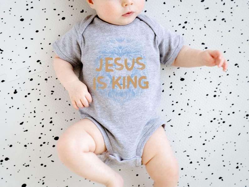 May include: A gray baby bodysuit with the text "JESUS IS KING" in gold and a light blue lion design. The baby is lying on a white surface with black speckles. The bodysuit is short-sleeved.