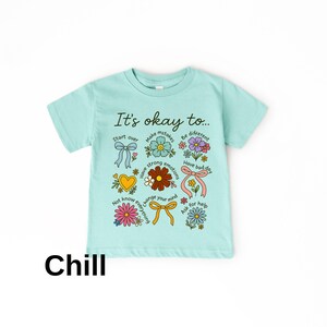 May include: A light blue t-shirt with the text "It's okay to..." and various floral and bow graphics. The shirt also has the word "Chill" printed at the bottom in black.