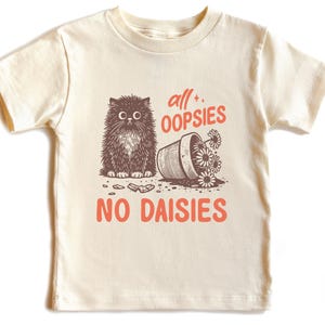 May include: A cream colored t-shirt with a black and white illustration of a cat knocking over a pot of flowers. The text "all oopsies no daisies" is printed in orange on the shirt.