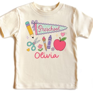 May include: A cream-colored short-sleeve t-shirt featuring a colorful graphic print of school supplies including a pencil, scissors, paintbrush, crayon, apple, and a pennant flag with the text Preschool. The name Olivia is printed in pink below.