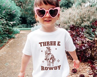 May include: White t-shirt with a brown graphic of a girl riding a horse. The text "THREE & ROWDY" is printed on the shirt.