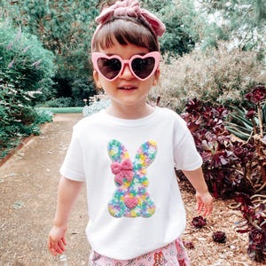 May include: A young child wearing a white t-shirt with a colorful bunny design and pink heart-shaped sunglasses. The bunny is made of a floral pattern with a pink bow. The child is standing outside on a path.