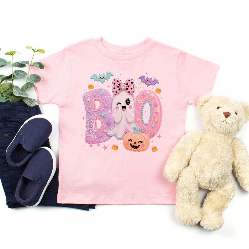Puede incluir: Una camiseta rosa claro con un estampado gr&aacute;fico de la palabra BOO en letras grandes de colores pastel. El dise&ntilde;o incluye un fantasma gui&ntilde;ando un ojo con un lazo de lunares rosa, una calabaza sonriente, murci&eacute;lagos peque&ntilde;os, estrellas y dulces.