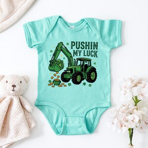 May include: A light teal baby bodysuit with a green tractor graphic. The tractor's bucket is filled with shamrocks, and gold coins are scattered around a pot of gold. Text reads "Pushin' My Luck."