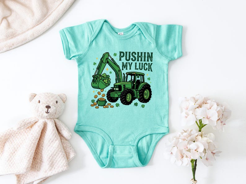 May include: A light teal baby bodysuit with a green tractor graphic. The tractor's bucket is filled with shamrocks, and gold coins are scattered around a pot of gold. Text reads "Pushin' My Luck."