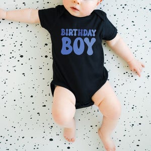 May include: A black baby bodysuit with the words "BIRTHDAY BOY" in blue. The baby is lying on a white surface with black splatters. The bodysuit has short sleeves and covers the baby's torso and legs.