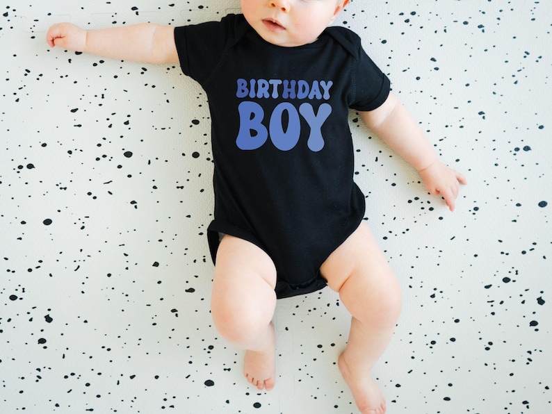 May include: A black baby bodysuit with the words "BIRTHDAY BOY" in blue. The baby is lying on a white surface with black splatters. The bodysuit has short sleeves and covers the baby's torso and legs.