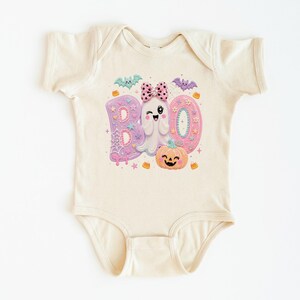 Puede incluir: Un body de manga corta color crema con un estampado gr&aacute;fico de la palabra BOO en letras rosa pastel y moradas. El dise&ntilde;o incluye un fantasma gui&ntilde;ando un ojo con un lazo rosa de lunares, una calabaza naranja sonriente, murci&eacute;lagos y estrellas.