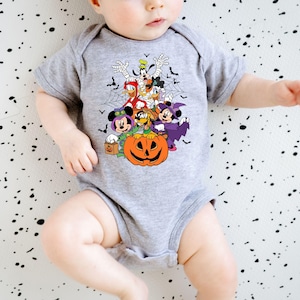 Op de afbeelding: Een grijze babybody met een Halloween-thema. Het ontwerp bevat stripfiguren zoals Mickey en Minnie Mouse, Donald Duck en Goofy, samen met een uitgesneden pompoen, vleermuizen en spinnenwebben. De body heeft korte mouwen.