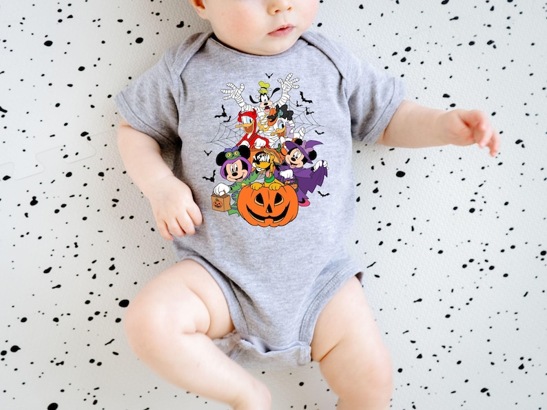 Op de afbeelding: Een grijze babybody met een Halloween-thema. Het ontwerp bevat stripfiguren zoals Mickey en Minnie Mouse, Donald Duck en Goofy, samen met een uitgesneden pompoen, vleermuizen en spinnenwebben. De body heeft korte mouwen.