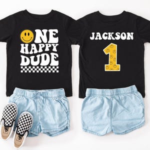 May include: Two black t-shirts with matching denim shorts and checkered slip-on sneakers. One shirt says "ONE HAPPY DUDE" with a smiley face. The other shirt says "JACKSON" and features a number one with smiley faces.
