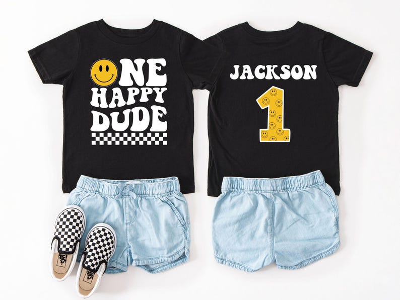 May include: Two black t-shirts with matching denim shorts and checkered slip-on sneakers. One shirt says "ONE HAPPY DUDE" with a smiley face. The other shirt says "JACKSON" and features a number one with smiley faces.
