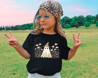 May include: A black t-shirt featuring a whimsical ghost wearing a witch hat, surrounded by daisies and foliage. The ghost has two small eye holes. The person in the image is wearing a leopard print scarf and sunglasses.