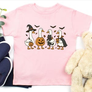 May include: Pink short-sleeved t-shirt featuring a Halloween-themed graphic. The graphic depicts four cartoon geese dressed in Halloween costumes: a witch, a jack-o'-lantern, a goose holding a black cat, and a vampire. Bats and leaves are also included.