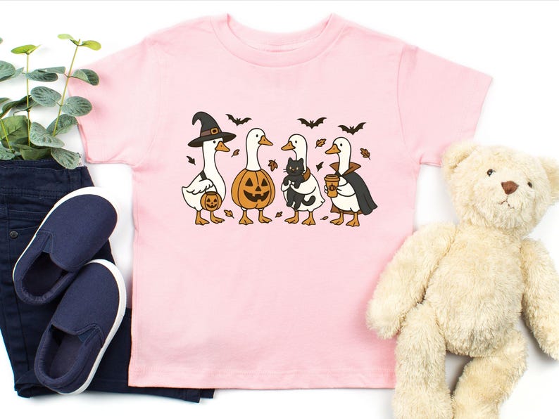 May include: Pink short-sleeved t-shirt featuring a Halloween-themed graphic. The graphic depicts four cartoon geese dressed in Halloween costumes: a witch, a jack-o'-lantern, a goose holding a black cat, and a vampire. Bats and leaves are also included.