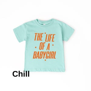 Pode incluir: Uma camiseta azul clara com o texto laranja "THE LIFE OF A BABY GIRL" e estrelas. A palavra "Chill" est&aacute; impressa em preto na parte inferior. Esta camiseta &eacute; projetada para beb&ecirc;s e crian&ccedil;as pequenas.
