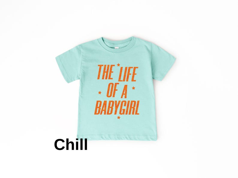 Pode incluir: Uma camiseta azul clara com o texto laranja "THE LIFE OF A BABY GIRL" e estrelas. A palavra "Chill" est&aacute; impressa em preto na parte inferior. Esta camiseta &eacute; projetada para beb&ecirc;s e crian&ccedil;as pequenas.