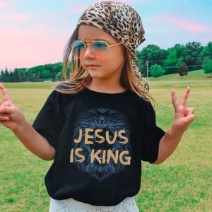 May include: Black t-shirt with the text "JESUS IS KING" in gold lettering and a lion's mane design. The person is wearing a leopard print scarf, sunglasses, and making a peace sign.