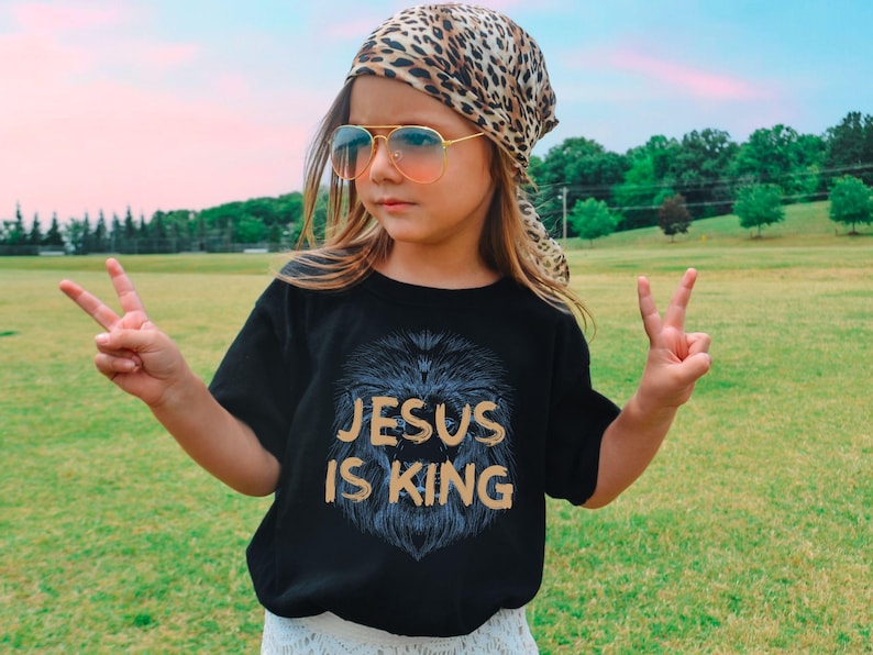 May include: Black t-shirt with the text "JESUS IS KING" in gold lettering and a lion's mane design. The person is wearing a leopard print scarf, sunglasses, and making a peace sign.