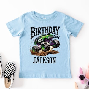 Pode incluir: Camiseta azul clara com um gr&aacute;fico de um monster truck verde saltando sobre terra, com o texto "BIRTHDAY BOY JACKSON". Sapatos slip-on xadrez em preto e branco tamb&eacute;m s&atilde;o vis&iacute;veis.
