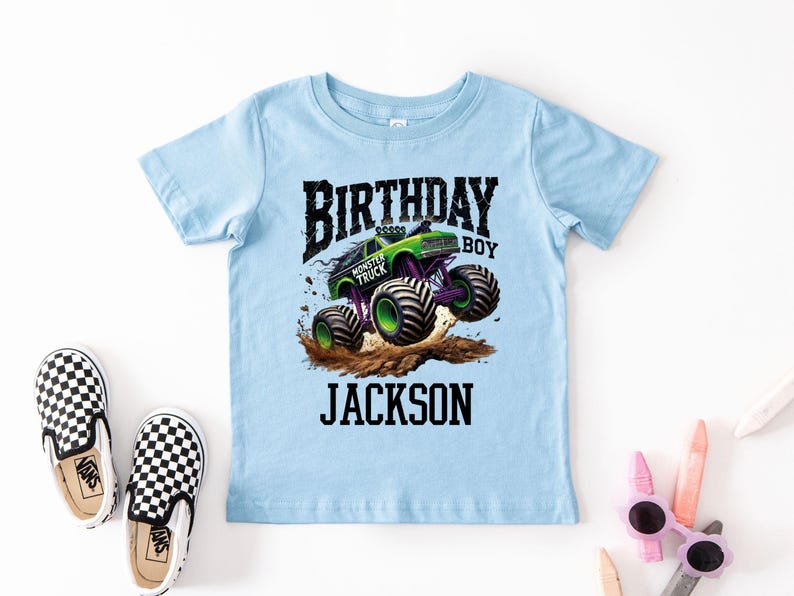 Pode incluir: Camiseta azul clara com um gr&aacute;fico de um monster truck verde saltando sobre terra, com o texto "BIRTHDAY BOY JACKSON". Sapatos slip-on xadrez em preto e branco tamb&eacute;m s&atilde;o vis&iacute;veis.
