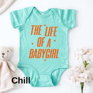 Pode incluir: Um body de beb&ecirc; turquesa com o texto "THE LIFE OF A BABYGIRL" em laranja. O texto &eacute; acentuado com pequenas estrelas laranja. Um ursinho de pel&uacute;cia e um cobertor branco est&atilde;o ao lado do body.