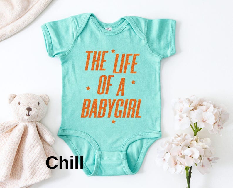 Pode incluir: Um body de beb&ecirc; turquesa com o texto "THE LIFE OF A BABYGIRL" em laranja. O texto &eacute; acentuado com pequenas estrelas laranja. Um ursinho de pel&uacute;cia e um cobertor branco est&atilde;o ao lado do body.