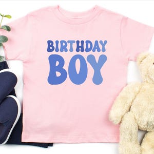 May include: A pale pink t-shirt with the words "BIRTHDAY BOY" in blue. The shirt is next to a pair of navy blue slip-on shoes, dark blue trousers, and a beige teddy bear.