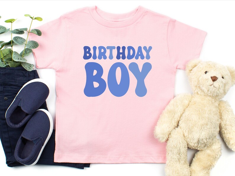 May include: A pale pink t-shirt with the words "BIRTHDAY BOY" in blue. The shirt is next to a pair of navy blue slip-on shoes, dark blue trousers, and a beige teddy bear.
