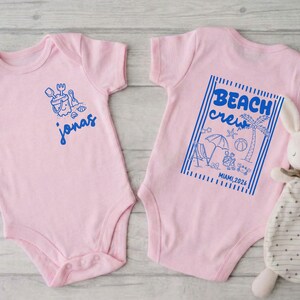 Op de afbeelding: Twee roze babyrompers. E&eacute;n heeft een blauwe afbeelding van een zandemmer en schep met de naam "Jonas". De andere heeft een blauwe afbeelding met de tekst "Beach Crew" met strandillustraties en "Miami 2026".