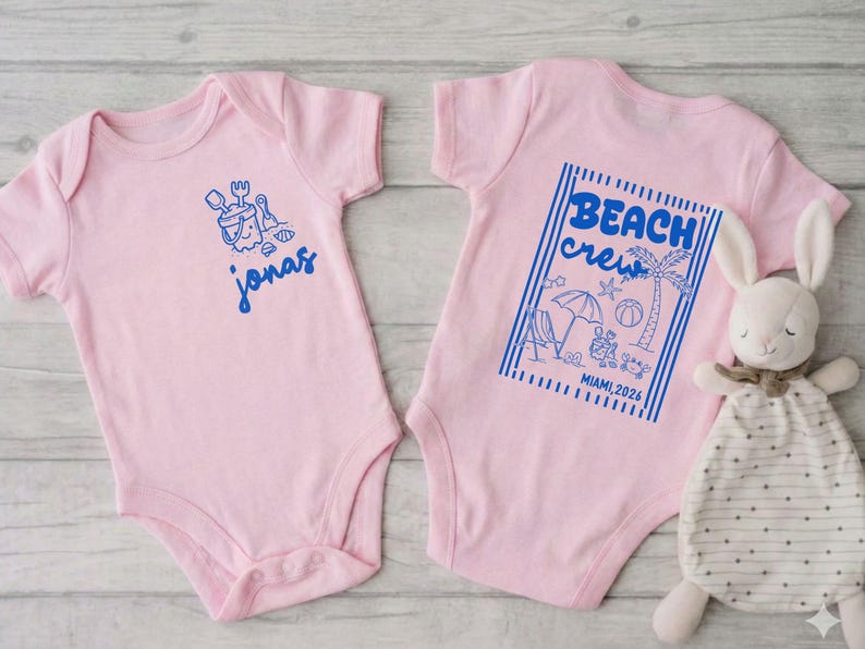 Op de afbeelding: Twee roze babyrompers. E&eacute;n heeft een blauwe afbeelding van een zandemmer en schep met de naam "Jonas". De andere heeft een blauwe afbeelding met de tekst "Beach Crew" met strandillustraties en "Miami 2026".