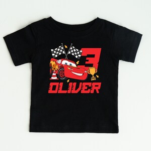 May include: Black toddler t-shirt featuring a red cartoon race car, the number 3, checkered flags, and a gold trophy. The name "OLIVER" is printed in red below the graphic. The shirt is designed for a child's birthday.