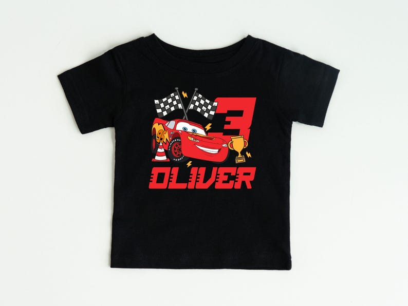 May include: Black toddler t-shirt featuring a red cartoon race car, the number 3, checkered flags, and a gold trophy. The name "OLIVER" is printed in red below the graphic. The shirt is designed for a child's birthday.