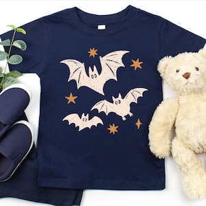 May include: A navy blue short-sleeve t-shirt featuring a graphic print of three light-colored bats and several orange stars. The bats are depicted in a stylized manner with simple facial features and spread wings across the center of the shirt.