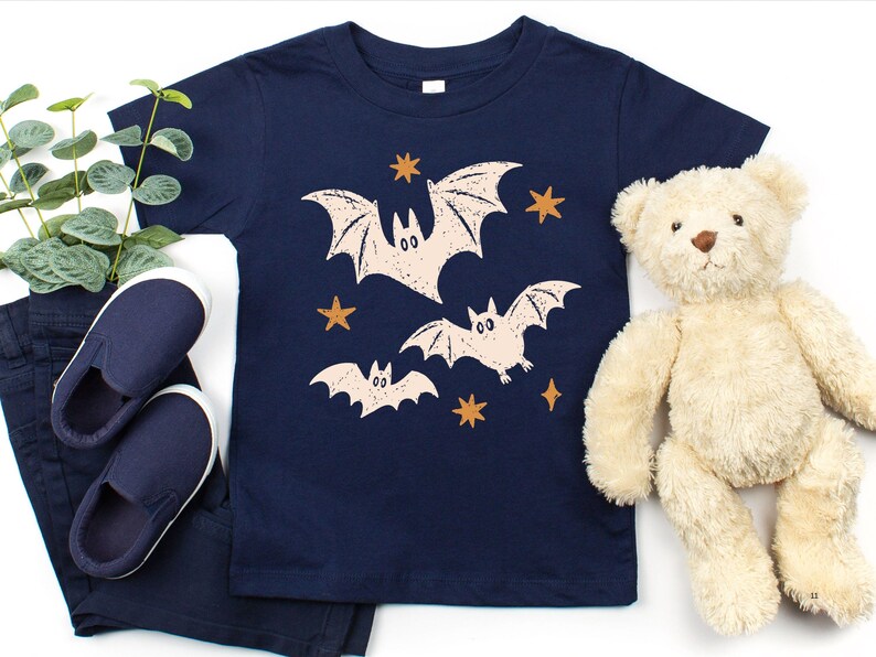 May include: A navy blue short-sleeve t-shirt featuring a graphic print of three light-colored bats and several orange stars. The bats are depicted in a stylized manner with simple facial features and spread wings across the center of the shirt.