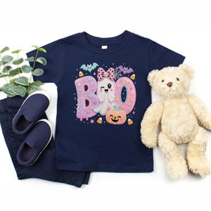 Puede incluir: Una camiseta azul marino de manga corta con un estampado gr&aacute;fico de la palabra BOO en rosa y morado pastel. El dise&ntilde;o incluye un fantasma gui&ntilde;ando un ojo con un lazo de lunares, una calabaza sonriente, murci&eacute;lagos, telara&ntilde;as y dulces.