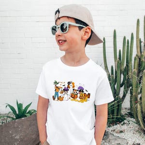 May include: A white t-shirt featuring a colorful Halloween-themed graphic. The graphic includes cartoon characters dressed in costumes, such as a ghost, a witch, and a pumpkin. The shirt is worn by a child wearing sunglasses and a baseball cap.