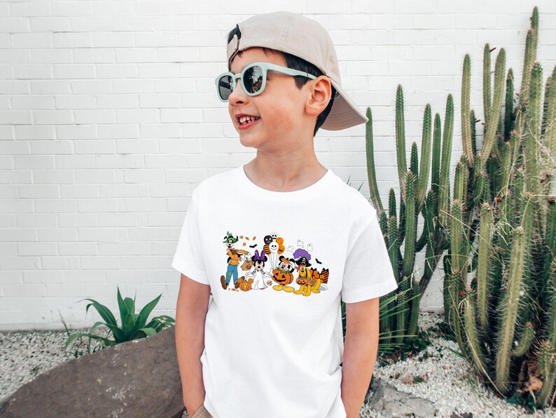 May include: A white t-shirt featuring a colorful Halloween-themed graphic. The graphic includes cartoon characters dressed in costumes, such as a ghost, a witch, and a pumpkin. The shirt is worn by a child wearing sunglasses and a baseball cap.