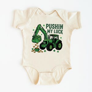 May include: Cream-colored baby bodysuit with a green excavator graphic scooping up shamrocks and gold coins. The text "PUSHIN MY LUCK" is printed above the excavator. A St. Patrick's Day themed baby outfit.