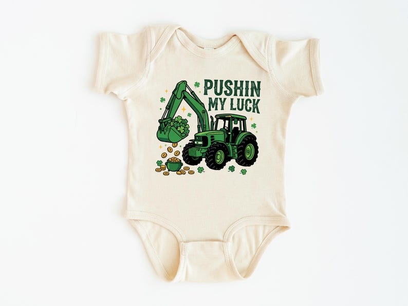 May include: Cream-colored baby bodysuit with a green excavator graphic scooping up shamrocks and gold coins. The text "PUSHIN MY LUCK" is printed above the excavator. A St. Patrick's Day themed baby outfit.